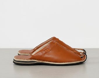 May include: A pair of brown leather slippers with a split toe design. The slippers have a stitched upper and a black and white striped sole. The footwear is designed for indoor use.