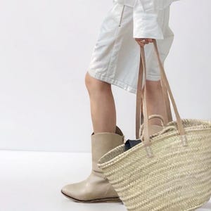 May include: A woven straw tote bag with tan leather handles. The bag is held by a person wearing a white outfit and tan boots. The bag has a rectangular shape and a natural color.
