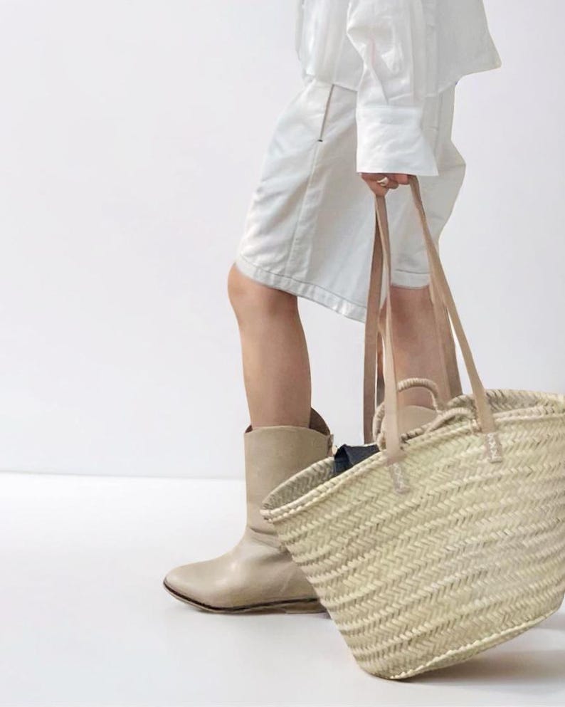 May include: A woven straw tote bag with tan leather handles. The bag is held by a person wearing a white outfit and tan boots. The bag has a rectangular shape and a natural color.