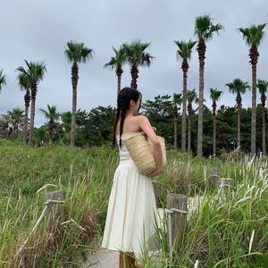 May include: A woman in a cream-colored dress and carrying a woven straw tote bag walks along a sandy path. Tall palm trees and green foliage create a tropical backdrop under an overcast sky. The dress is strapless and the tote bag is beige.