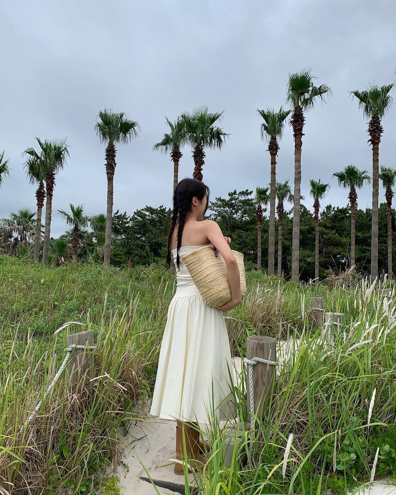 May include: A woman in a cream-colored dress and carrying a woven straw tote bag walks along a sandy path. Tall palm trees and green foliage create a tropical backdrop under an overcast sky. The dress is strapless and the tote bag is beige.