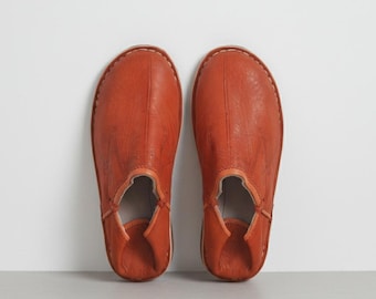 May include: A pair of orange leather slippers. The slippers have a rounded toe and a low-cut design around the ankle. The leather has a textured appearance, and the stitching is visible around the edges. The slippers are displayed on a light gray surface against a white background.