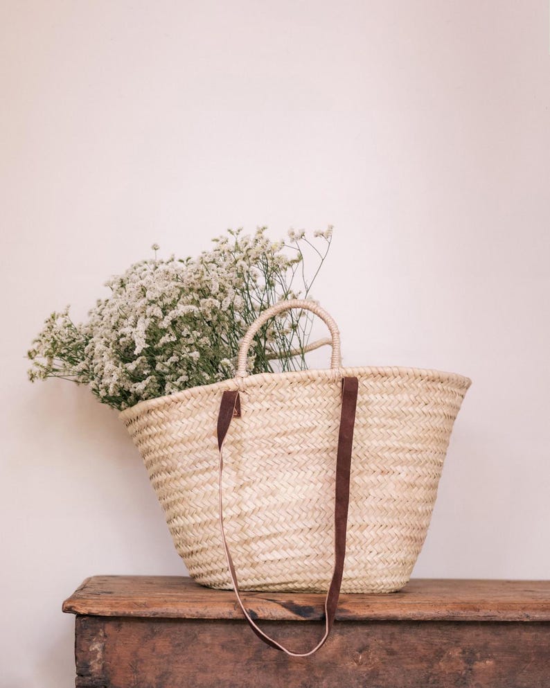 May include: A woven straw tote bag with brown leather straps and handles, filled with white flowers. The bag is sitting on a rustic wooden surface, with a light pink background.
