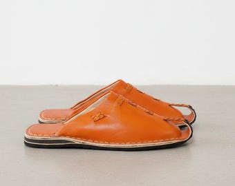 May include: A pair of orange leather slippers with a stitched design. The slippers have a black and white sole and a simple, open-toe design. The slippers are made of a soft, supple leather.