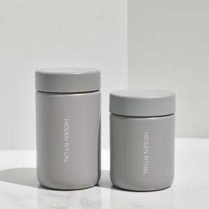 May include: Two gray cylindrical containers with the text "HIDDEN RITUAL" printed on the front.