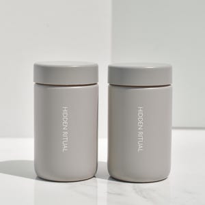 May include: Two gray cylindrical containers with the text "HIDDEN RITUAL" printed on the front.
