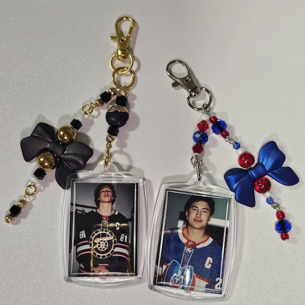 Könnte beinhalten: Zwei Hockey-Schlüsselanhänger. Einer hat eine schwarze Schleife, goldene Perlen und ein Foto eines Hockeyspielers. Der andere hat eine blaue Schleife, rote und blaue Perlen und ein Foto eines Hockeyspielers. Beide haben goldene und silberne Verschlüsse.