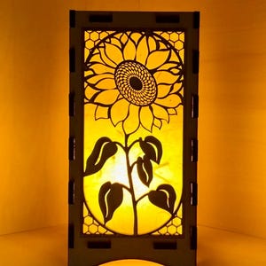 May include: A wooden lamp with a cutout design of a sunflower. The lamp is lit and casts a warm glow.