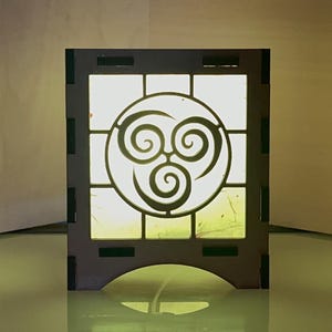 Puede incluir: Una lámpara de madera con un acabado marrón claro y un panel translúcido blanco con un símbolo negro estilizado de tres espirales entrelazadas. La lámpara tiene una pequeña base con un arco redondeado.