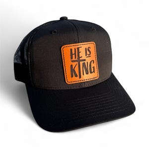 May include: Black trucker hat with a mesh back and a curved brim. The hat features a square, orange leather patch with the words "HE IS KING" in white lettering and a cross symbol.