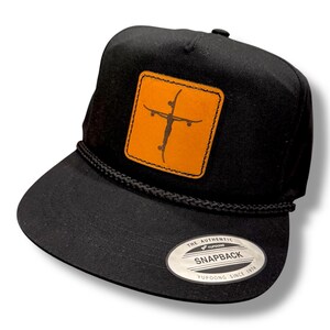 May include: Black snapback hat with a square, tan leather patch featuring an orange skateboard cross design. The hat has a flat brim, a black rope detail, and a silver tag that reads "Snapback".
