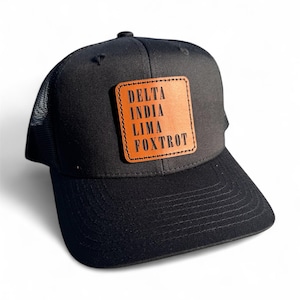 May include: Black trucker hat with a mesh back and a curved brim. A square, tan leather patch on the front displays the text "DELTA INDIA LIMA FOXTROT" in black capital letters. The hat is set against a white background.
