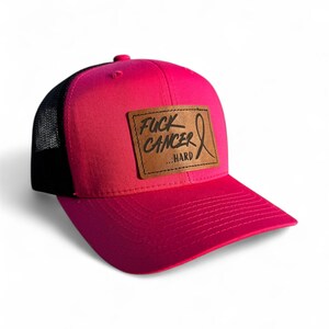 May include: A pink trucker hat with a black mesh back. The front of the hat features a brown leather patch with the words "FUCK CANCER...HARD" and a pink ribbon graphic.