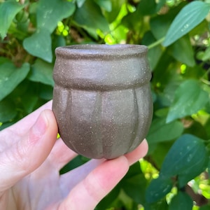 May include: A small, dark brown ceramic cup with a speckled texture and a rounded, bulbous shape. The exterior features vertical indentations around the lower body and a smooth, slightly flared rim, creating a distinct, tactile surface finish.