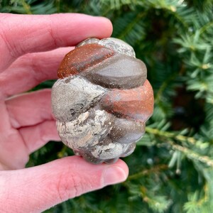 May include: A hand-held, rounded stone sculpture with a textured surface. The stone features a mix of colors, including shades of gray, brown, and reddish-brown. The sculpture's form is organic and appears to be a natural, hand-carved piece.