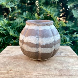 May include: A small, brown ceramic vase with a pumpkin-like shape. The vase features horizontal bands of light tan and dark brown. The vase is sitting on a wooden surface, with green foliage in the background. This decorative vase is suitable for small plants or flowers.