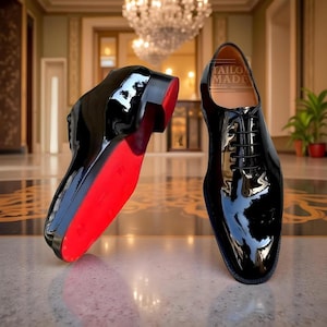 May include: Two patent leather black dress shoes with red soles. One shoe is upright, the other is tilted, revealing the red sole. The shoes feature a classic lace-up design. The text "TAILOR MADE SHOES" is visible inside one shoe.