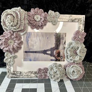 May include: A white picture frame with a distressed finish is decorated with crocheted flowers in shades of purple and gray. The frame holds a photo of the Eiffel Tower in Paris.