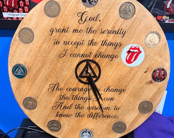May include: A round wooden wall clock featuring the Serenity Prayer in black script text. The clock face is adorned with twelve circular recovery medallions placed at each hour marker and a red and white tongue logo sticker on the right side.