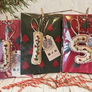 May include: Three felt Christmas ornaments shaped like the letters "J" and "S", and a rectangular shape, each with floral embroidery and red berry accents. They are packaged in cellophane with decorative tags and hung with twine. The background features red and green paper with Christmas tree cutouts.