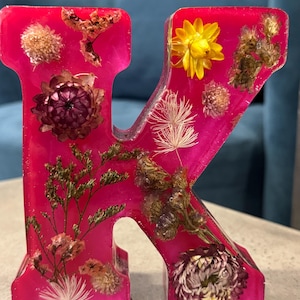 May include: A hot pink resin letter "K" with dried flowers embedded within. The flowers are various colors, including yellow, purple, and white. The letter is standing upright on a light gray surface.