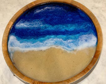 May include: A round wooden tray with a resin design depicting a beach scene. The tray features a sandy shore at the bottom, transitioning to foamy white waves and deep blue ocean. The wooden rim adds a natural touch.