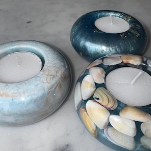 May include: Three tealight candle holders. One is light blue with a textured surface, another is a deep blue with gold flecks, and the third is blue with seashells embedded in the resin. Each holds a white tealight candle.