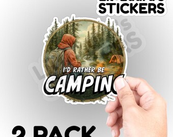 May include: A 2-pack of stickers featuring a camping scene with the text "I'd Rather Be Camping." The stickers have a circular design with a person in a red hoodie and a tent in the background. The brand name "Lil Laura's STICKERS" is at the top.