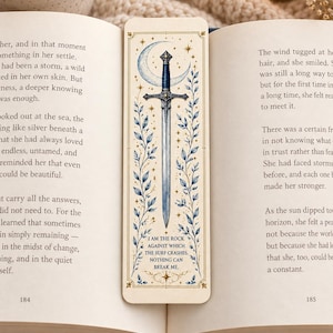 May include: A rectangular paper bookmark featuring a central illustration of a silver sword with a blue hilt, set against a crescent moon and gold stars. Blue leafy vines frame the sword. The text reads: I AM THE ROCK AGAINST WHICH THE SURF CRASHES. NOTHING CAN BREAK ME.