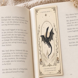 May include: A rectangular paper bookmark featuring a black ink illustration of a dragon in flight above mountain peaks, enclosed in an oval frame with floral accents. The text reads Fourth Wing at the top and A dragon without its rider is a tragedy. A rider without their dragon is dead at the bottom.