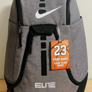 May include: Gray backpack with black accents, a white Nike logo, and a basketball-themed tag. The tag displays the number 23, with space for a name and team name. The word "ELITE" is printed on the front.