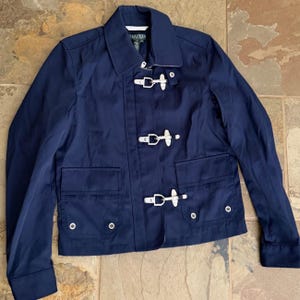 May include: A navy blue jacket featuring a pointed collar, long sleeves, and a front closure with three silver-toned metal toggle clasps. The jacket includes two front flap pockets with silver-toned grommet details and a Lauren Ralph Lauren label.
