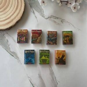 Op de afbeelding: Een verzameling van zeven miniatuur Harry Potter-boeken gerangschikt in twee rijen. Elk boek toont de iconische omslagillustratie en titeltekst van J.K. Rowling, waaronder Steen der Wijzen, Geheime Kamer, Gevangene van Azkaban, Vuurbeker, Orde van de Feniks, Halfbloed Prins en Relieken van de Dood.
