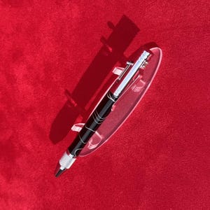May include: A black and silver pen with a decorative design, displayed on a clear stand against a red background. The pen features a silver clip and tip, with a patterned black barrel. The pen is designed for writing.