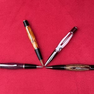 May include: Four handcrafted pens with unique designs are arranged on a red background. The pens feature various colors, including wood grain, white, and black accents. The pens are positioned to converge at a central point.
