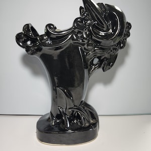 May include: A glossy black ceramic vase with an ornate, sculptural design. The vase features a flared top with intricate details, a slender neck, and a rounded base. The overall shape is reminiscent of a stylized mythical creature.