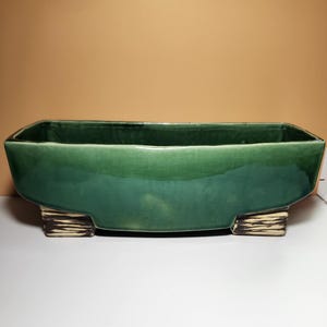 May include: A rectangular, emerald green ceramic planter with a glossy finish. The planter is supported by two rectangular, textured, light brown and white feet. The planter is designed for indoor or outdoor use.