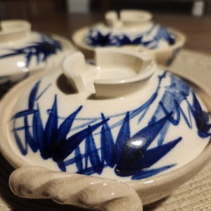 May include: A set of three ceramic bowls with lids. The lids are off-white with a blue leaf pattern. Each bowl has a twisted handle. The bowls are on a woven mat.