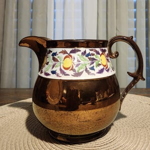 May include: A decorative brown pitcher with a gold base and handle. The pitcher features a white band with a floral design in yellow, purple, green, and blue. The pitcher is likely made of ceramic or porcelain.