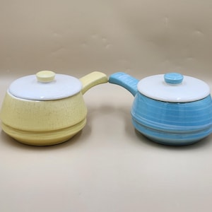 May include: Two ceramic saucepans with lids, one yellow with a crackle glaze and one blue with horizontal ridges. Each pot features a white lid with a small knob handle and a side handle extending from the main body of the cookware.