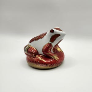 May include: A ceramic frog figurine with a white head and red and gold accents. The frog is sitting on a red base with gold detailing. The figurine has a glossy finish and is likely a decorative item.