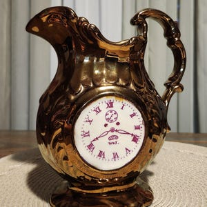 May include: A gold-colored decorative pitcher with a clock face. The clock face has Roman numerals and is surrounded by a white background. The pitcher has a handle and a spout.
