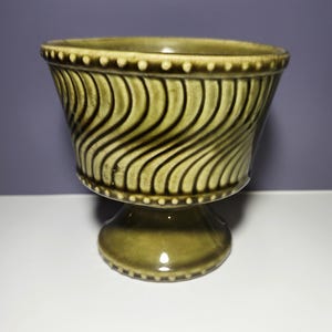 May include: A green ceramic pedestal bowl with a flared, textured body. The bowl features a wavy pattern and a beaded rim. The base is a rounded pedestal. The bowl is likely vintage and suitable for decorative use.
