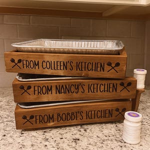 Op de afbeelding: Houten serveerschalen met drie niveaus en aluminium bakken. Elke schaal is gegraveerd met "From Colleen's Kitchen", "From Nancy's Kitchen" en "From Bobbi's Kitchen" in het zwart. De schalen staan op een granieten aanrecht.
