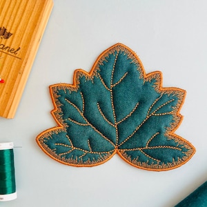 Pode incluir: Aplique em forma de folha verde-azulado com costuras laranja. O aplique está sobre uma superfície azul clara, junto a carretéis de linha verde e um porta-alfinetes de madeira com o texto "City mimo manel".