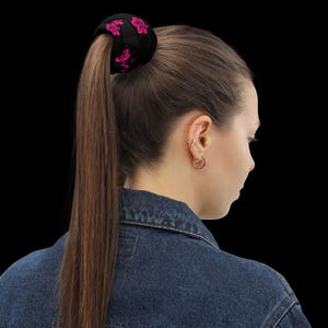 May include: A black fabric hair scrunchie featuring a repeating pattern of bright pink hibiscus flowers. The scrunchie is secured around a long, straight brown ponytail, contrasting with the dark background and the blue denim jacket collar.