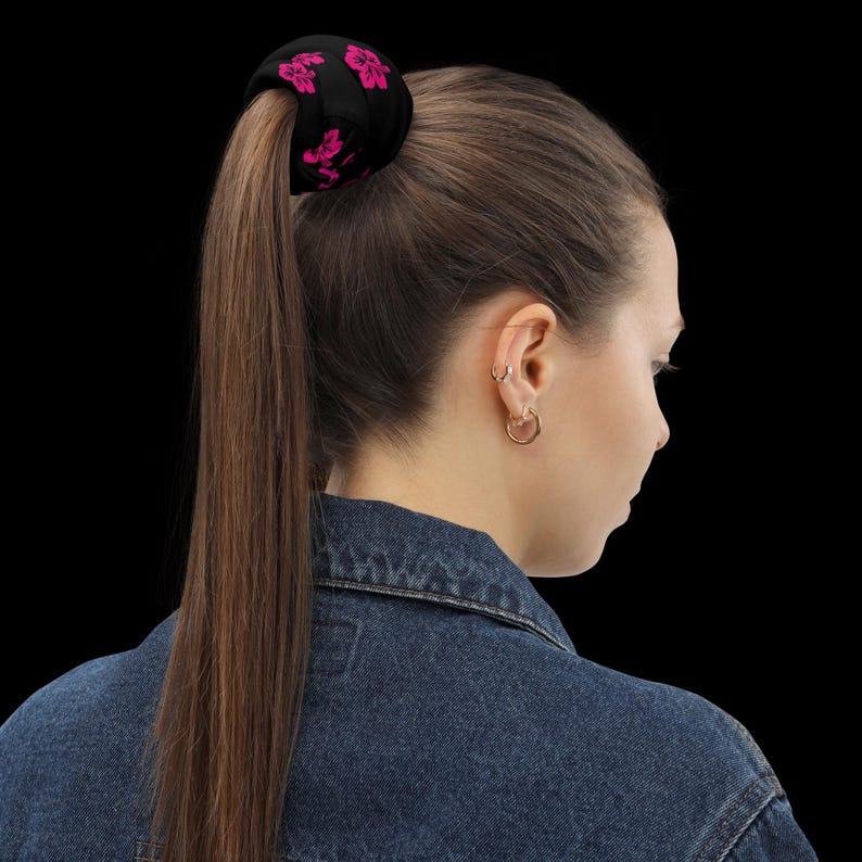 May include: A black fabric hair scrunchie featuring a repeating pattern of bright pink hibiscus flowers. The scrunchie is secured around a long, straight brown ponytail, contrasting with the dark background and the blue denim jacket collar.