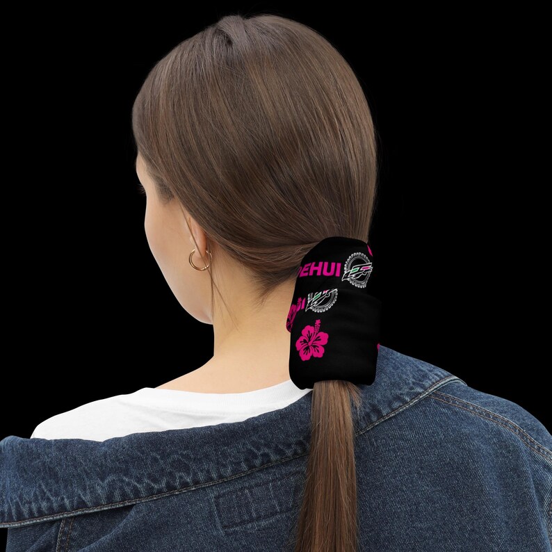 May include: A black hair scrunchie featuring a repeating pattern of bright pink hibiscus flowers and the white text "DEHUI" alongside a circular graphic logo. The accessory is wrapped around a ponytail of brown hair, securing the hair in place.