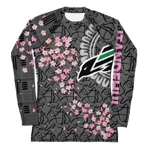 May include: A long-sleeve athletic shirt featuring a dark grey background with a black abstract line pattern. The design includes pink cherry blossom branches, a stylised black and green bird graphic, and the text ITANDEHII in pink on the sleeve.