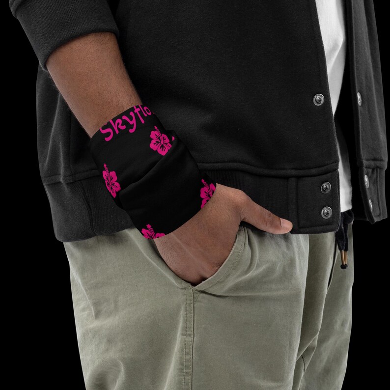 May include: A black neck gaiter featuring a repeating pattern of bright pink hibiscus flowers and the text Skyflo in a matching pink script font. The fabric is worn around the wrist, displaying the floral design and the printed text clearly.